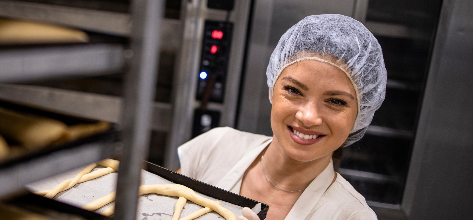Women smiling at the camera while in the commercial kitchen, holding a tray of unbaked goods and wearing a Bouffant hair net.